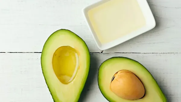 An avocado cut in half and filled with oil in the side without the pip stands next to a small bowl of oil.