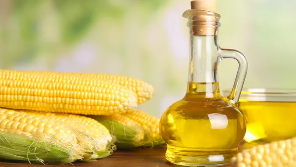 A glass bottle of oil stands next to dehusked corn cobs.