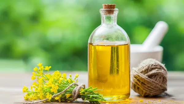 A bottle of oil with a cork stopper stands on a wooden table next to a bunch of canola flowers tied with string.