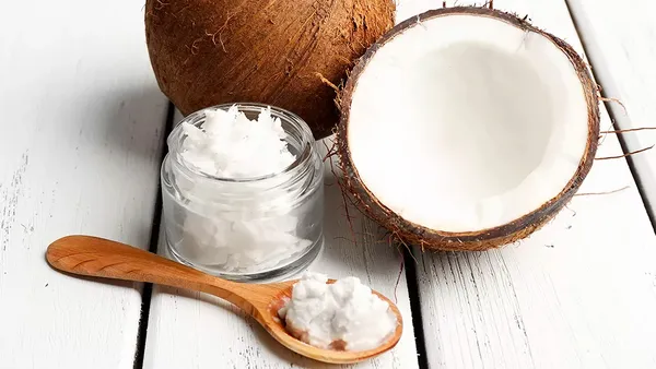 A half a coconut stands next to a small glass jar and a wooden spoon filled with coconut meat.