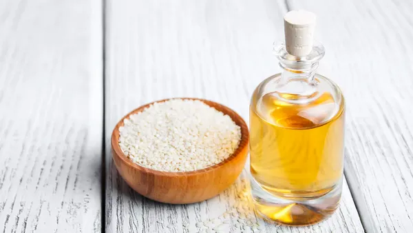 A small wooden bowl of white sesame stands next to a glass bottle of oil.