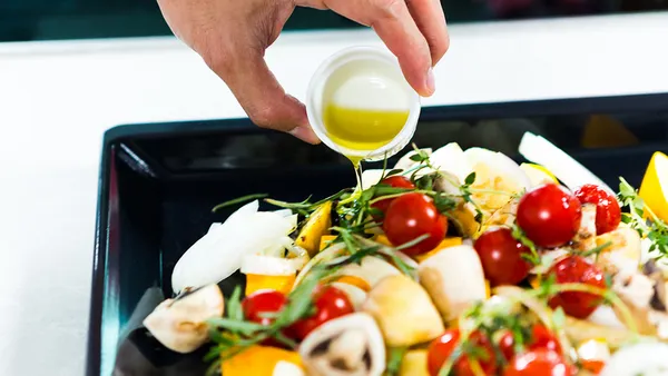 A hand drizzles oil over fresh vegetables in a roasting pan.