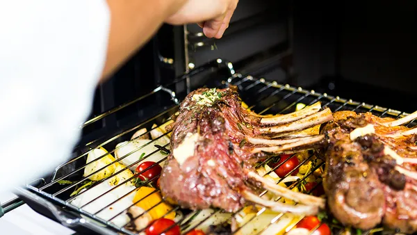 A hand with tongs reaches to turn chops on a grill rack over a pan of vegetables.