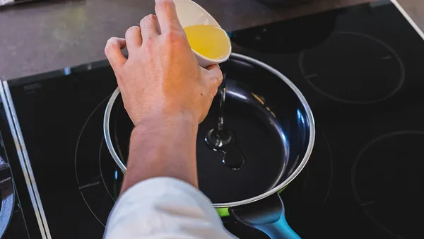 A hand pours oil from a small bowl into a skillet.