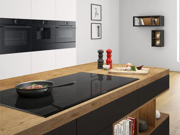 A modern kitchen containing a kitchen island with a wooden counter and an integrated hob with a frying pan on it. In the background three dark appliances are integrated into a white cabinet.
