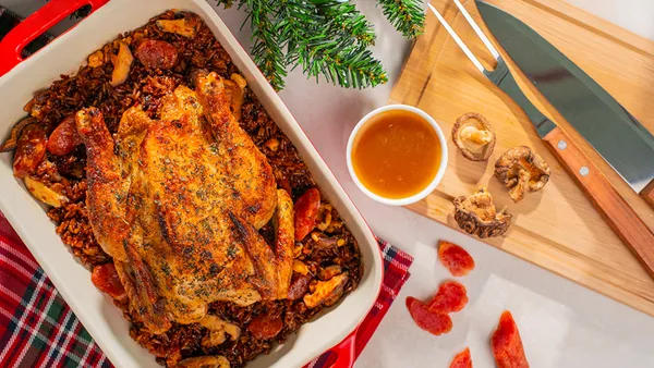 A casserole dish containing a whole roast chicken on a bed of peppers stands next to a wooden chopping board with a carving knife and fork.