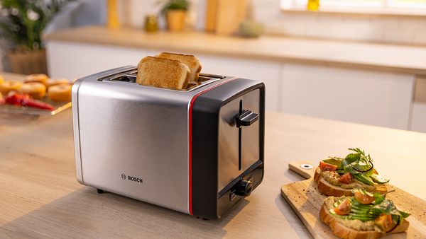 Nerezový toaster s plátkami toastov vedľa dosky s toastami obloženými lososom a avokádom na kuchynskej linke.