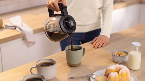 Woman pouring coffee from glass jug into mug placed on kitchen counter.