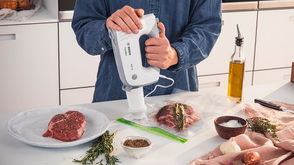 Eine Person arbeitet an einer Arbeitsplatte, umgeben von Olivenöl, Steaks, Zwiebeln und Kräutern, und verwendet einen Bosch Handrührer mit dem Vakuumierzubehör, um Beutel mit Zutaten für Sous-Vide luftdicht zu verschließen.