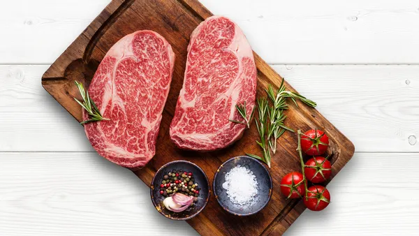 Two raw steaks on a wooden board with small plates of spices and tomatoes.