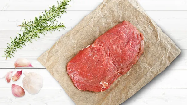 A piece of firm, fresh red meat on a piece of brown paper with rosemary and whole garlic cloves.