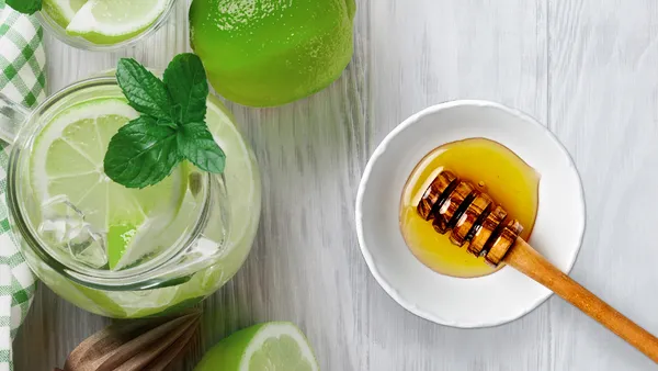 A view from above of a glass of a fresh lime drink with a sprig of mint in it and limes as well as a little honey next to it.