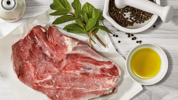 Ein Stück frisches rotes Fleisch, daneben eine Schüssel mit Öl, schwarzer Pfeffer im Mörser und Minzzweige.