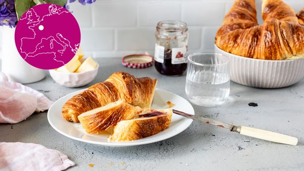 Eine winzige Karte von Frankreich überlagert die Aufnahme einer Küchenarbeitsplatte mit einem Teller, auf dem ein soeben mit Marmelade bestrichenes Croissant liegt.   