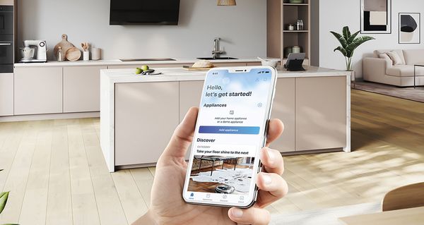 Ένα χέρι το οποίο κρατάει ένα smartphone σε πρώτο πλάνο, με μια όμορφη εικόνα κουζίνας στο φόντο.