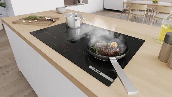 A downdraft induction hob with a steak sizzling in a frying pan next to boiling pot containing vegetables.