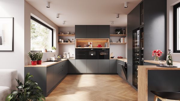 Cocina moderna con elegantes armarios oscuros, encimeras de madera y luz natural procedente de una gran ventana, con plantas y una decoración elegante.