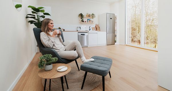 Een meisje zit in een stoel en geniet ontspannen van een kopje koffie, met op de achtergrond een keuken met een groot raam.