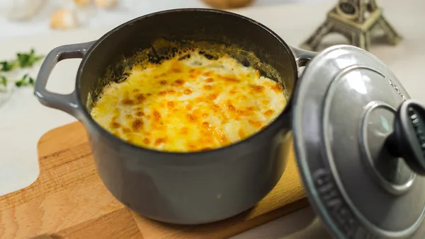 A cast iron pot containing a freshly baked celeriac dauphinoise stands on a wooden board with its lid resting on its side.