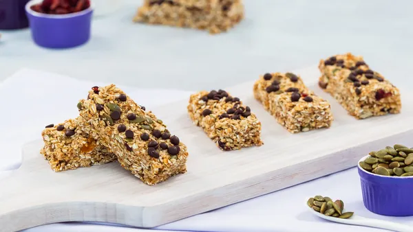  Five granola bars are lined up on a white wooden board and surrounded by ingredients like nuts and dried fruit. 