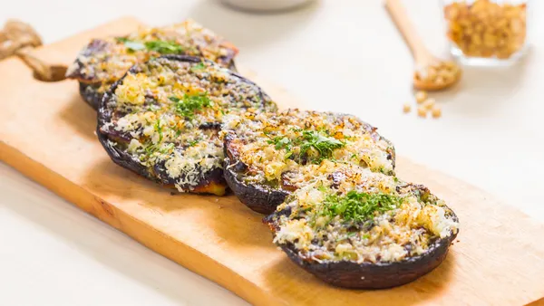 Halloumi and pesto-stuffed mushrooms on a wooden board next to glass bowl filled with herbs. 