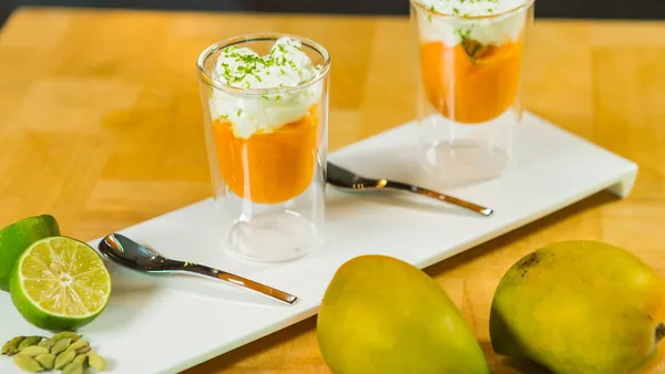  Two glasses filled with a mango, cardamom and whipped cream dessert stand on a white platter behind two fresh mangoes. 