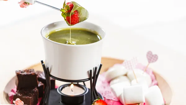  A fork with a coated strawberry hovers above a bowl of matcha fondue in a stand above a lit candle. 