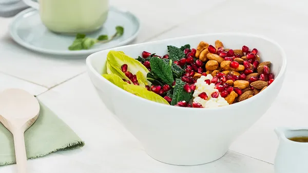 A pomegranate, nut and kale salad in a white bowl stands next to a jug of dressing and wooden spoon on a green napkin. 