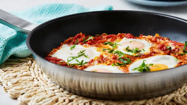 A pan with shakshuka and eggs rests on a woven mat. 