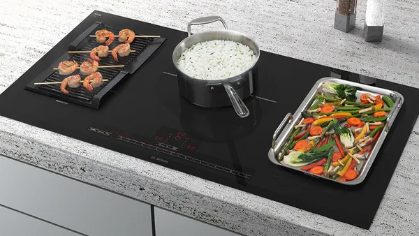 A kitchen countertop with an integrated induction cooktop featuring teppanyaki with vegetables, a grill plate with prawn skewers, and a pot of rice in the center.