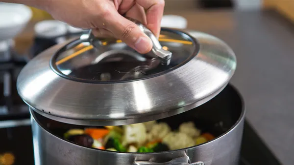 A person is lifting the lid of a cooking pot filled with vegetables.