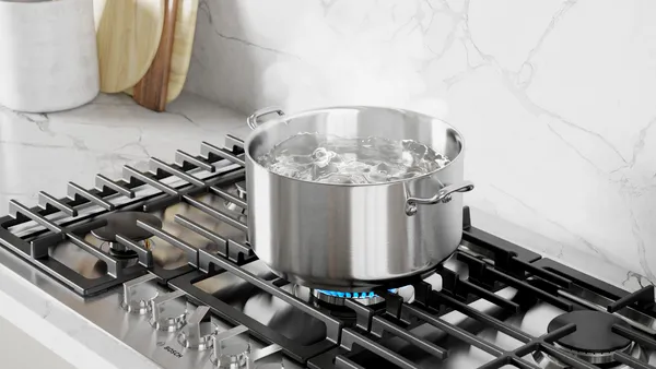 A stainless steel cooking pot with boiling water on a gas stove in a bright kitchen environment.