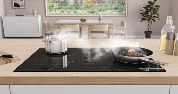 Boschin keittotaso integroidun liesituuletinmoduulin kanssa. Pannu, jolla on pihvi, ja kattila keittotasolla, taustalla ruokailualue ja ruokapöytä.