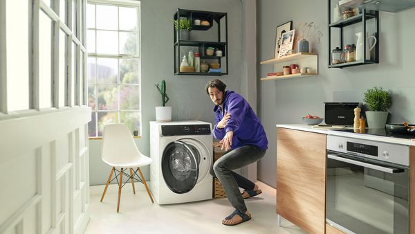 Muž v malé kuchyni nadšeně ukazuje na svou nově pořízenou pračku se sušičkou Serie 8. Spotřebič slouží jako provizorní stůl, na jehož desce je úhledně prostřená snídaně s ubrusem.