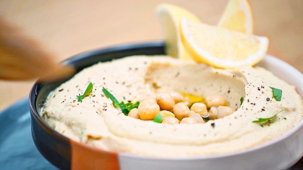 Hummus angerichtet mit Zitronenschnitzen, Kräutern, Kichererbsen und Olivenöl in einem Keramikschälchen.