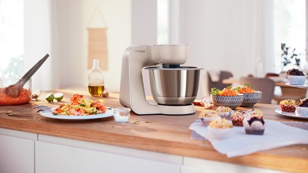 Eine Bosch K&uuml;chenmaschine auf einer Arbeitsplatte umringt von Salat, Pasta und Muffins.