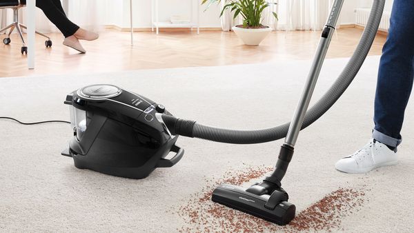 Eine Person saugt mit einem Staubsauger ohne Beutel von Bosch der Serie 8 Schmutz von einem hellen Teppichboden in einem modernen Wohnzimmer.