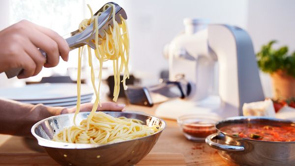 Frisch zubereitete Pasta wird mit einer Zange aus einer Metallschüssel entnommen.