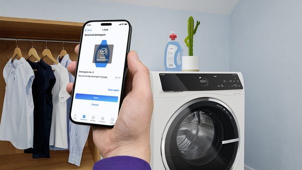 Hånd holder en smartphone med den åbne Home Connect-app foran en vaske-/tørremaskine med en flaske vaskemiddel og en kaktus med tommelfingrene opad oven på. 