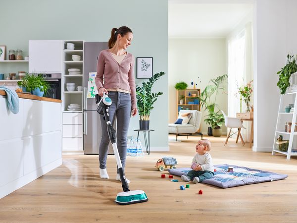 Noor ema pühib kööki Unlimited 7 ProHygienic Aquaga. Tema kõrval mängib väike laps puitpõrandal tekil väikeste mänguasjadega.