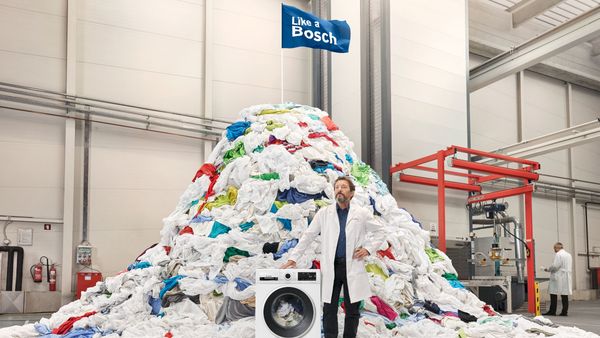 Mann steht vor einem 10 Tonnen großen Wäscheberg neben einer Bosch Waschmaschine. Bosch Waschmaschinen durchlaufen 10 Tonnen Wäschen in Qualitäts- und Testzentren, bevor sie auf den Markt kommen.