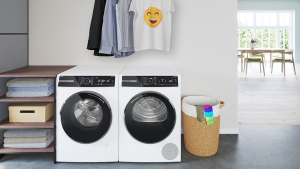 Ein Bosch Waschmaschinen- und Trockner-Set steht nebeneinander, darüber hängen mehrere Kleidungsstücke auf einer Stange; daneben ein Korb mit bunter Wäsche in einem hellen Waschraum.