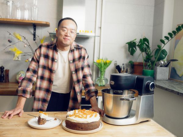 En person ikl&auml;dd flanellskjorta st&aring;r vid disken bredvid en matberedare med en kaka toppad med apelsinskivor och en kakbit p&aring; en tallrik.