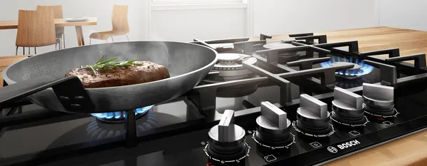 In a bright white kitchen, there is a kitchen island with a built-in induction cooktop displaying teppanyaki with prawns and a grill plate with meat. In the background, there are kitchen cabinets with built-in ovens and a dishwasher.