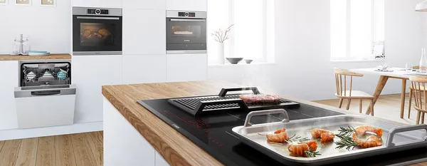 In a bright white kitchen, there is a kitchen island with a built-in induction cooktop displaying teppanyaki with prawns and a grill plate with meat. In the background, there are kitchen cabinets with built-in ovens and a dishwasher.