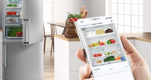 Une main tient un smartphone montrant l'intérieur d'un réfrigérateur, visible dans la cuisine lumineuse à l'arrière-plan.