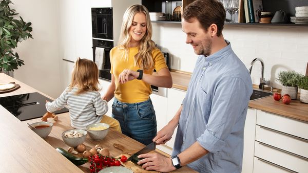 Un jeune couple et son enfant en bas âge se trouvent dans une cuisine moderne et lumineuse. L'homme coupe des légumes, tandis que la femme regarde sa montre connectée.