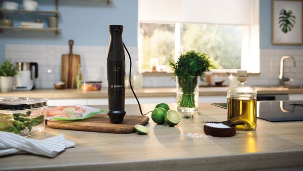 Frullatore a immersione Bosch appoggiato su un tagliere in una cucina luminosa, circondato da lime, olio, sale e verdure