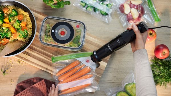 Piano di lavoro con frullatore a immersione Bosch che mette sottovuoto un sacchetto di carote, accanto a un recipiente con verdure