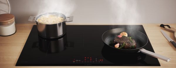 Une table à induction Bosch avec une casserole d’asperges et un steak dans une poêle.